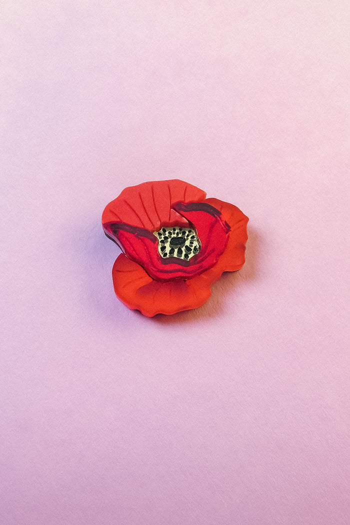 A realistic handmade Red Poppy Flower Lapel Pin with a yellow and black center is placed on a soft, light pink surface, making it a charming accessory for fashion enthusiasts.