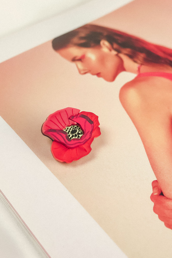 A Red Poppy Flower Lapel Pin brooch rests on an open magazine page featuring a woman with slicked-back hair in a pink top, set against a soft pink background—perfect for fashion enthusiasts seeking a chic, handmade touch.