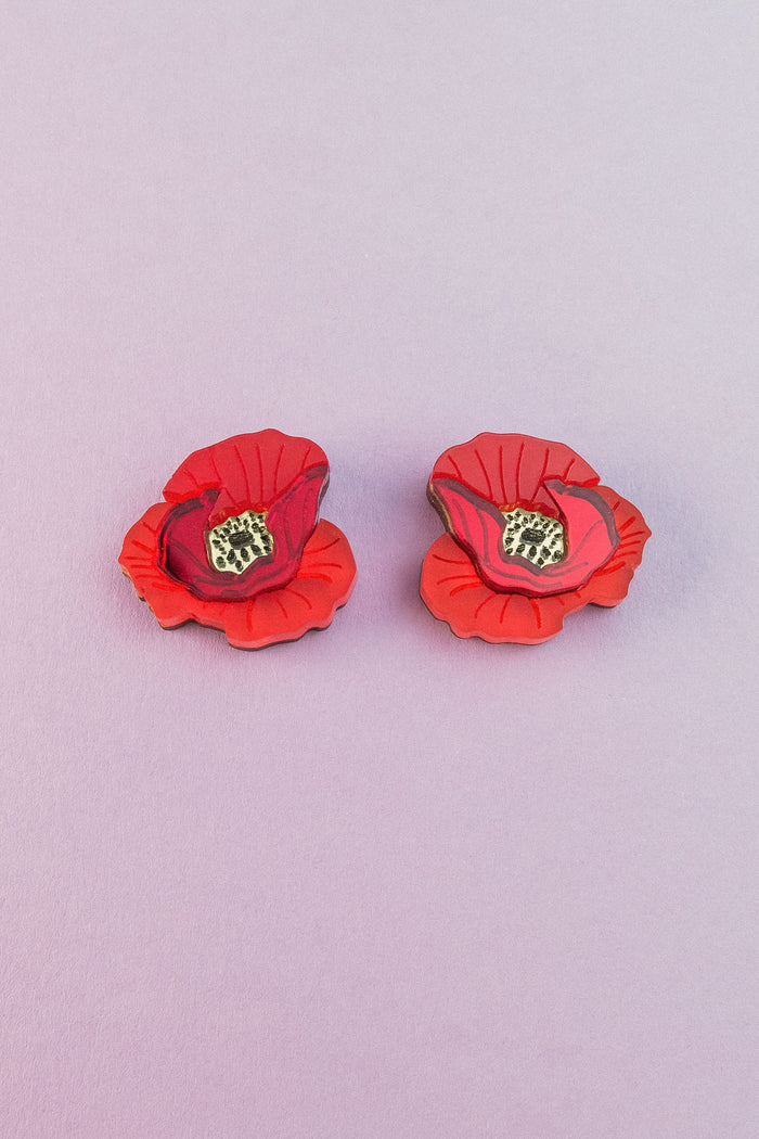 Two lightweight acrylic earrings featuring a vibrant poppy design with black and yellow centers are displayed on a light purple background.