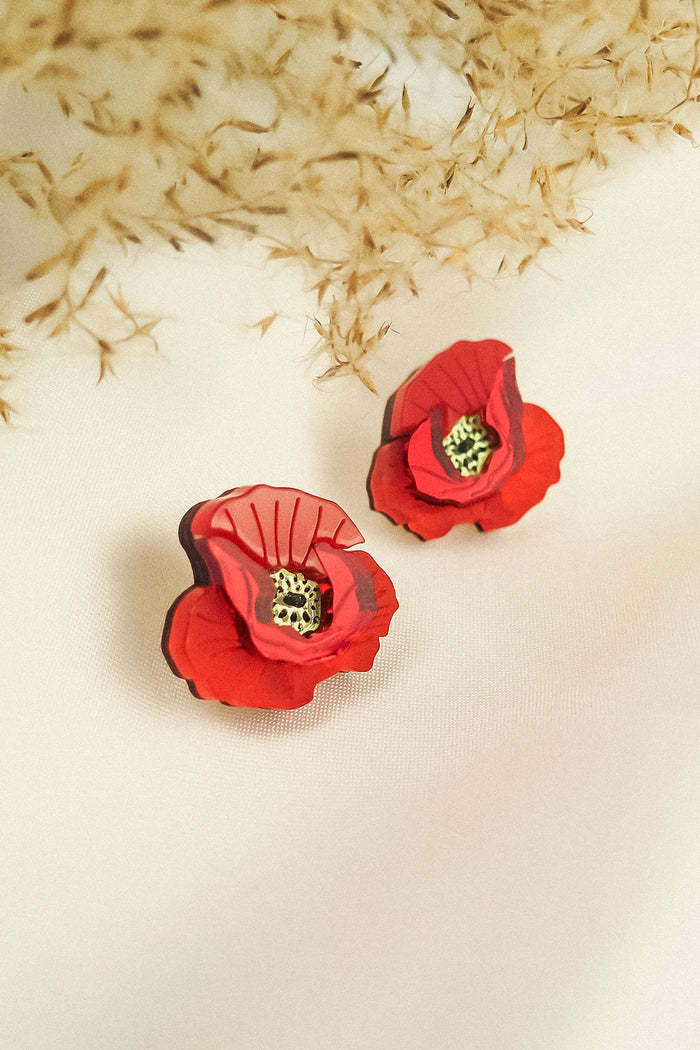 Two red poppy design acrylic earrings with black and yellow centers rest on a white fabric surface. Beige dried foliage in the background creates a soft, elegant setting, highlighting their unique style and lightweight feel.