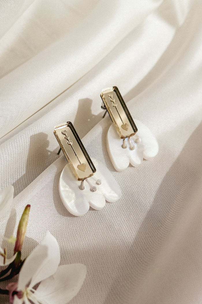 A pair of handcrafted earrings with gold rectangular studs engraved with small designs, attached to white, petal-shaped drops, displayed on a soft, cream-colored fabric with a white flower nearby.