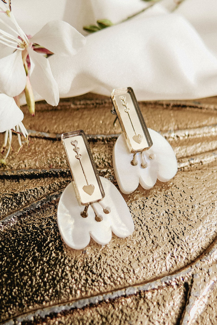 Rectangular gold earrings with engraved key and heart designs rest on white shell-like bases, displayed on a textured metallic surface with white flowers nearby, offering allergy-free comfort.