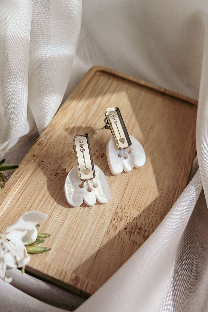 Two pairs of elegant earrings, including Romantic Tulip Earrings with pearl-like white and gold elements, are displayed on a light wooden tray amid soft fabric and blooms, offering allergy-free comfort in a delicate, inviting scene.