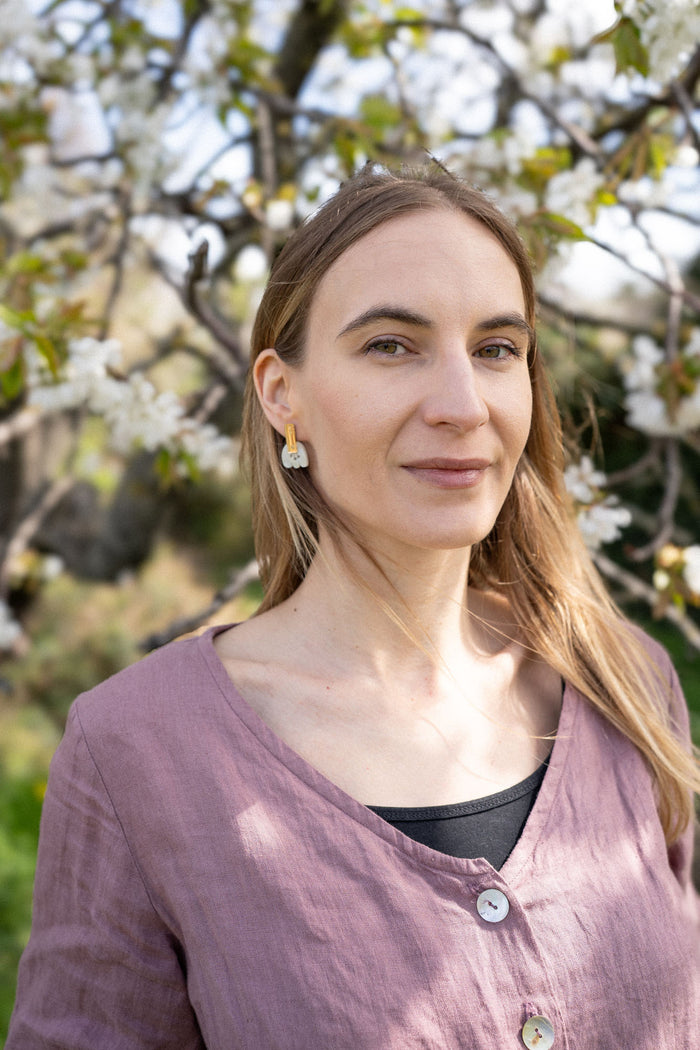 A woman with long blonde hair, wearing a mauve top and Romantic Tulip Earrings in marbled purple acrylic, stands outdoors in front of a blooming tree with white flowers. Sunlight highlights her face and the spring blossoms behind her.