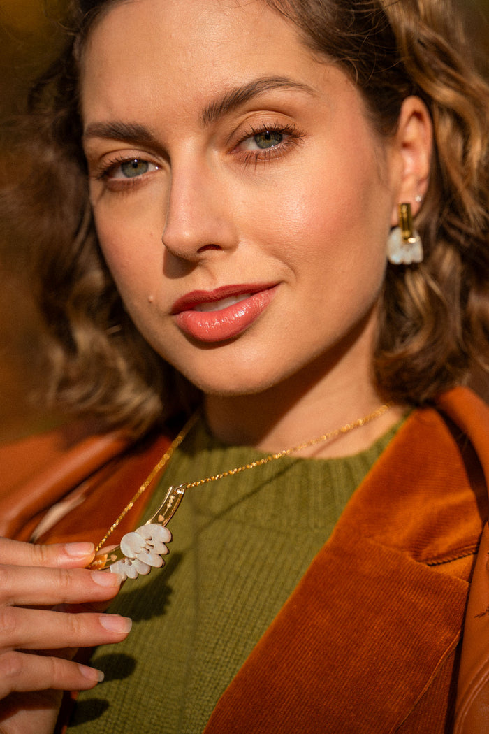 A woman with wavy brown hair wears an elegant green top, brown jacket, and handcrafted gold jewelry with matching white flower-shaped earrings and a Tulip Necklace, holding the necklace and looking confidently at the camera.