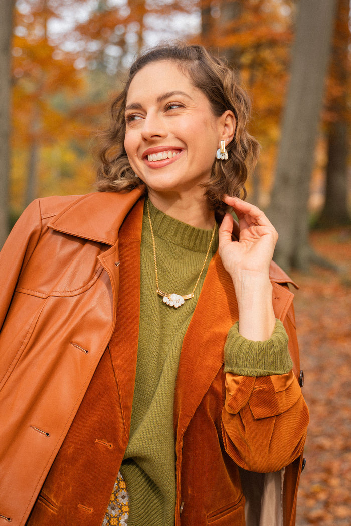 A woman smiles elegantly while standing in a forest with autumn leaves. She wears a brown leather coat over an orange suede jacket, with a green sweater, patterned skirt, and a handcrafted Tulip Necklace. Tall trees and fallen leaves fill the background.