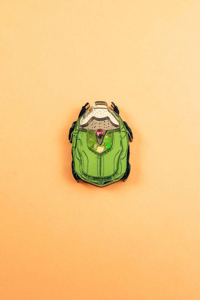 A green and black enamel Rose Chafer Pin, resembling a sci-fi or robot helmet, is centered on a plain peach background—a meaningful design perfect for lovers of handcrafted accessories.