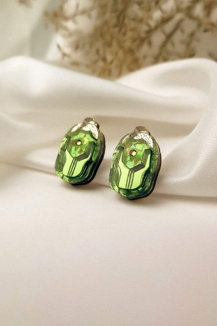 A pair of green, faceted gemstone Rose Chafer Studs with metallic tops displayed on white satin fabric, these nature-inspired earrings shine against a soft, blurred background.