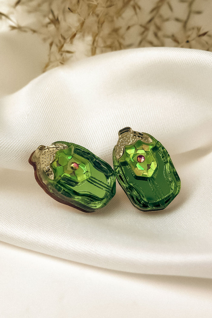 Two green, oval-shaped glass craft beads with a faceted surface and a small pink center gem, perfect for nature-inspired earrings, resting on a white silky fabric backdrop with blurred dried flowers in the background.