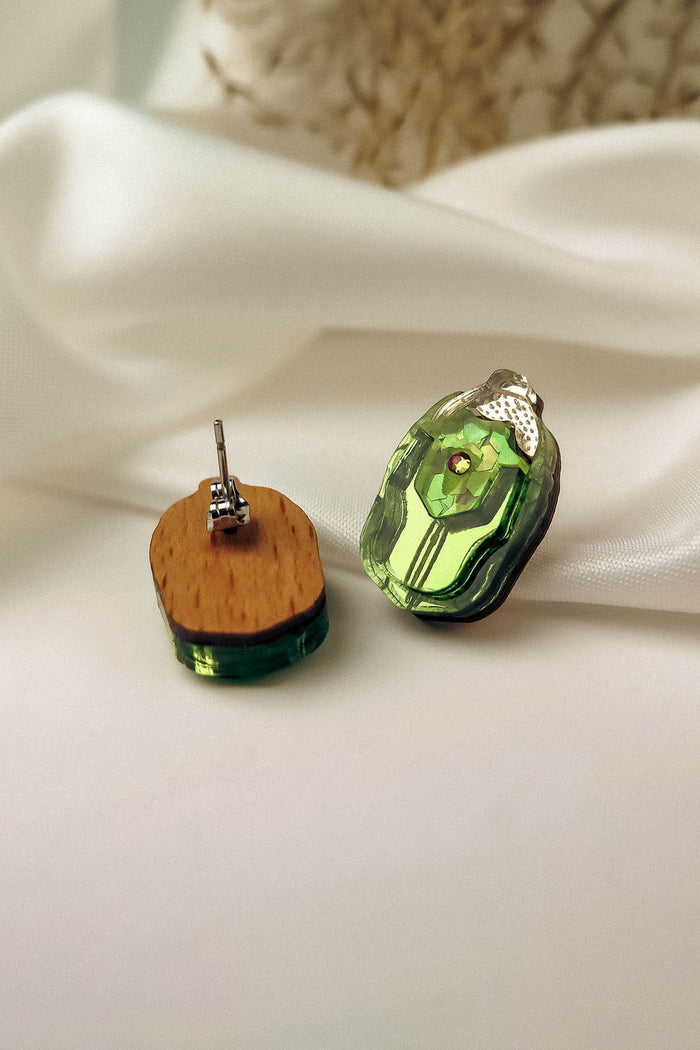 A pair of handcrafted Rose Chafer Studs with green translucent stones and wooden backs are displayed on white satin fabric. One nature-inspired earring shows the front, while the other reveals the wooden back and metal post.