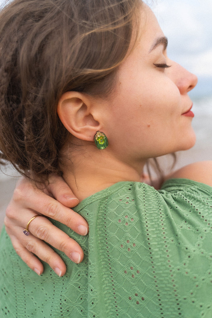 A woman with closed eyes and a peaceful expression rests her hand on her shoulder. She wears a green, eyelet-patterned top and handcrafted earrings. Her brown hair is slightly tousled as she stands outdoors.