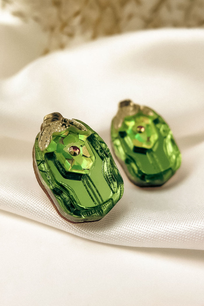 A close-up of Rose Chafer Studs, handcrafted earrings featuring green gemstones with gold accents, resting on soft white satin. Their shiny, faceted surface and decorative top make them perfect for nature lovers.