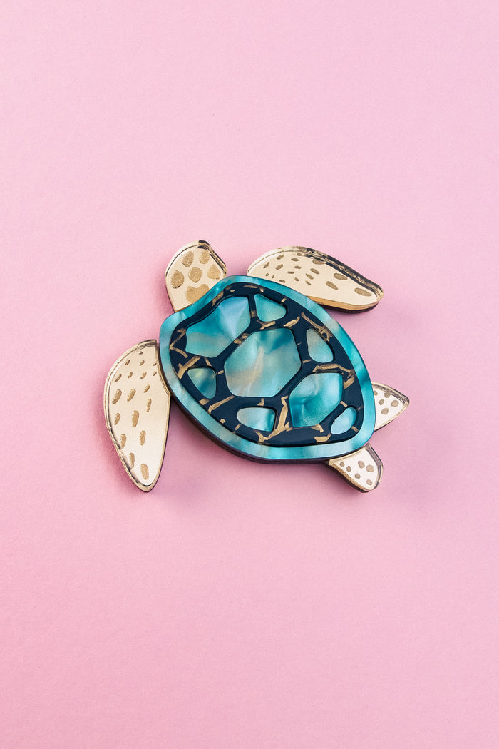 A handcrafted sea turtle brooch featuring an artistic design with a blue and teal shell and beige, patterned limbs, placed on a light pink background.