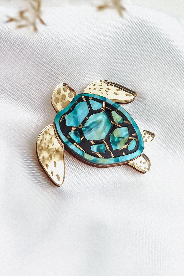 A handcrafted sea turtle brooch with gold-toned limbs and a turquoise, blue, and black artistic design on its shell, displayed on a white fabric background.