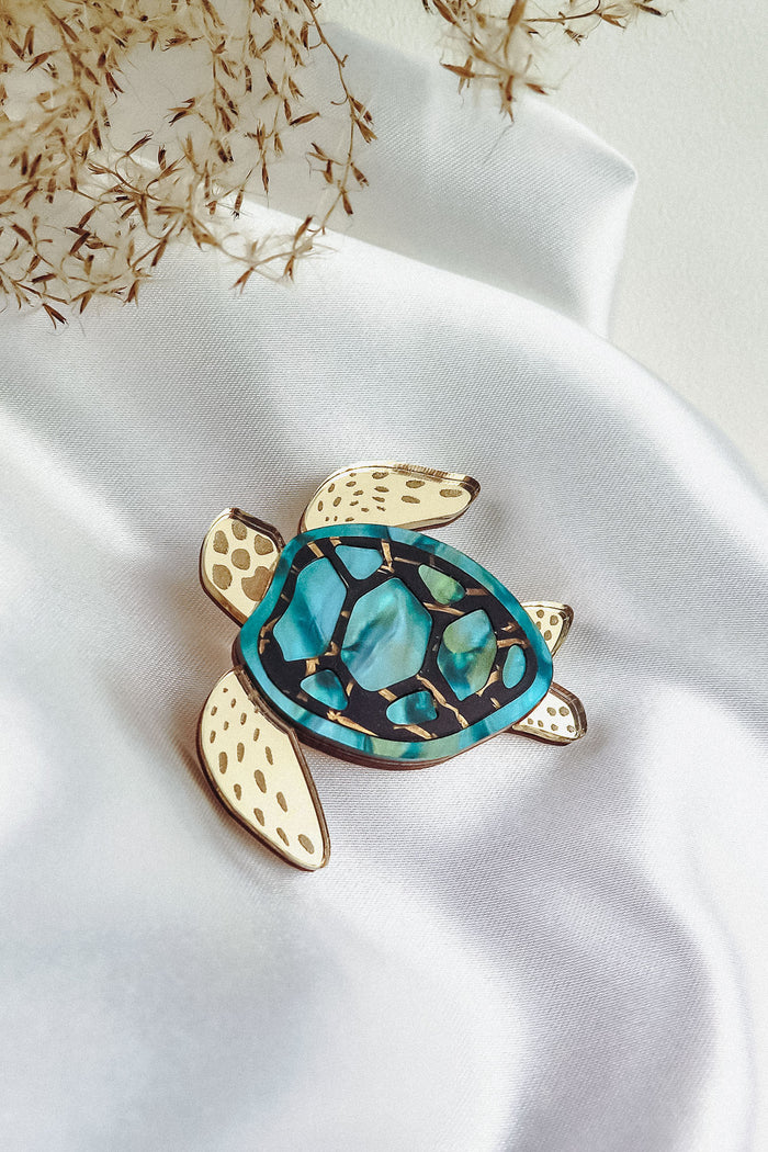 A handcrafted sea turtle brooch with a gold outline, cream-colored flippers, and a blue and green mosaic-patterned shell is displayed on white satin fabric with dried grasses nearby.