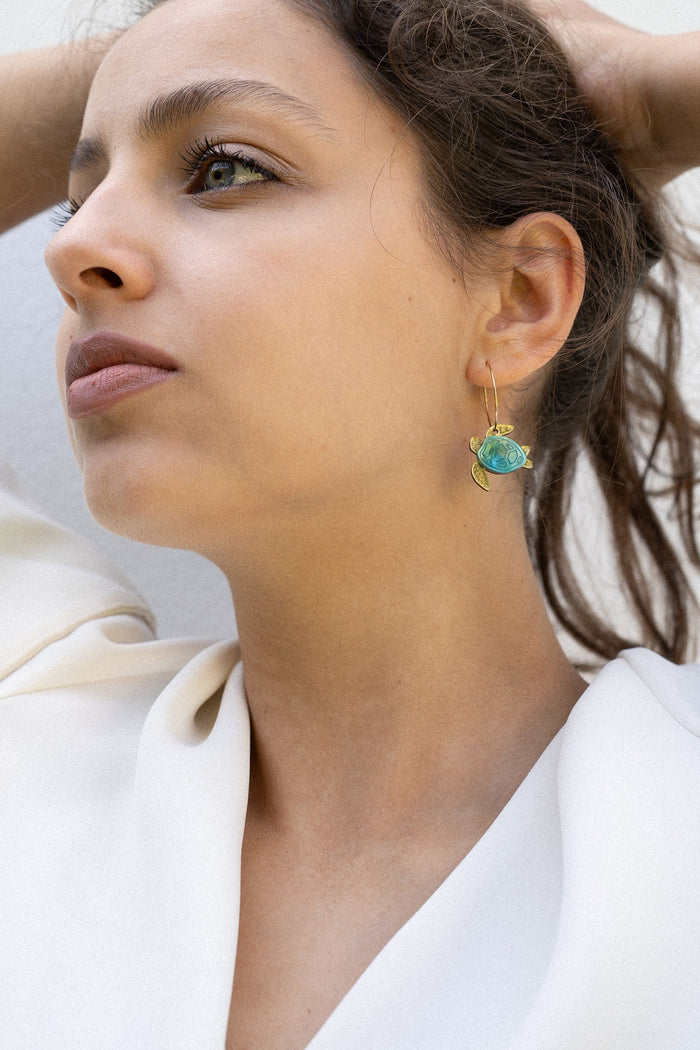 A woman with dark hair pulled back holds her hands behind her head, wearing a white blouse and handcrafted Little Turtle Hoop Earrings with green gemstones, looking thoughtfully into the distance against a plain light background.