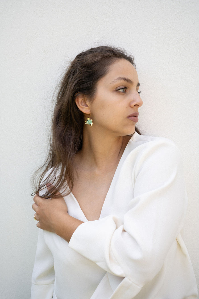A woman with long brown hair wearing a white top and handcrafted Little Turtle Hoop Earrings looks to her right, standing against a plain white wall and gently holding her hair with one hand.