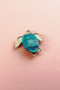A small sea turtle pin, handcrafted with gold-colored flippers and a blue mirror-acrylic glass shell, rests on a light pink background.