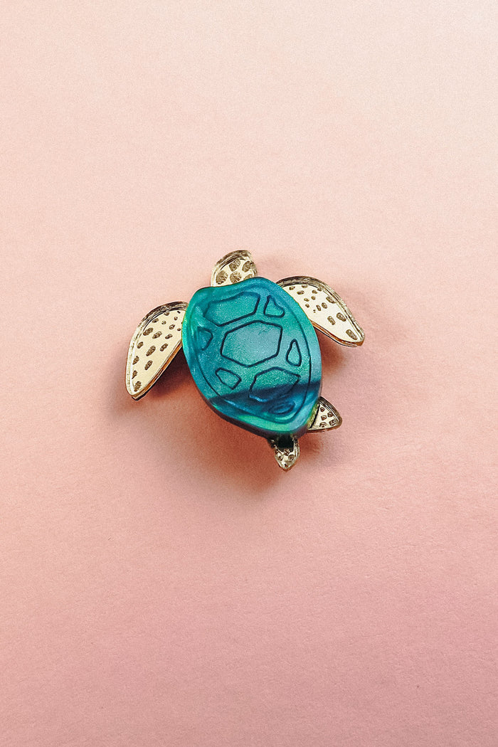 A small sea turtle pin, handcrafted with gold-colored flippers and a blue mirror-acrylic glass shell, rests on a light pink background.
