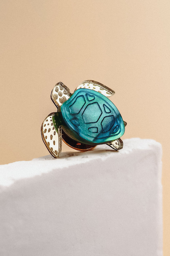 A handcrafted sea turtle pin with a blue-green shell and gold-toned, textured flippers is perched on the edge of a white textured surface against a beige background.