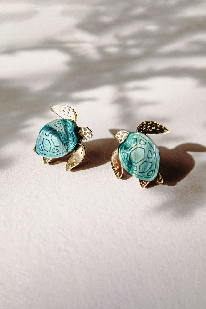 Two small Sea Turtle Studs with turquoise shells and gold-colored heads, legs, and tails rest on a light surface, casting soft shadows in natural light for a summery vibe.