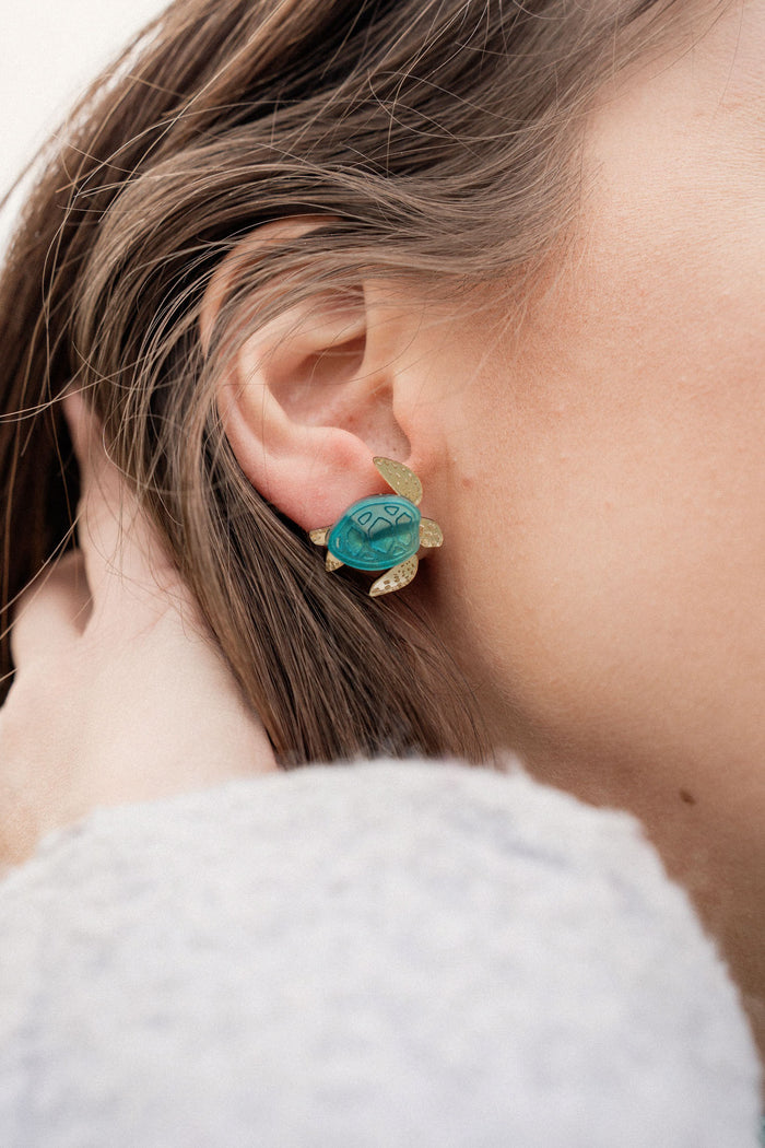 A close-up of a persons ear wearing gold and turquoise Sea Turtle Studs, with their light brown hair partially visible, captures an ocean-inspired, summery vibe.