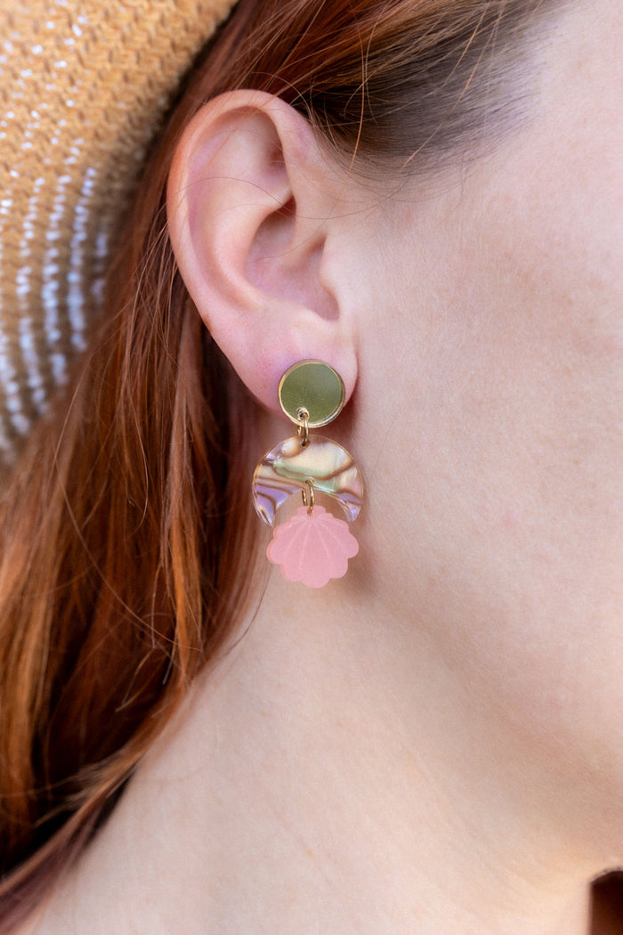 A close-up of a womans ear wearing a handmade gold stud earring with a bow and a dangling pink seashell-shaped charm. She has red hair, is wearing a straw hat, and the lightweight design adds delicate charm.