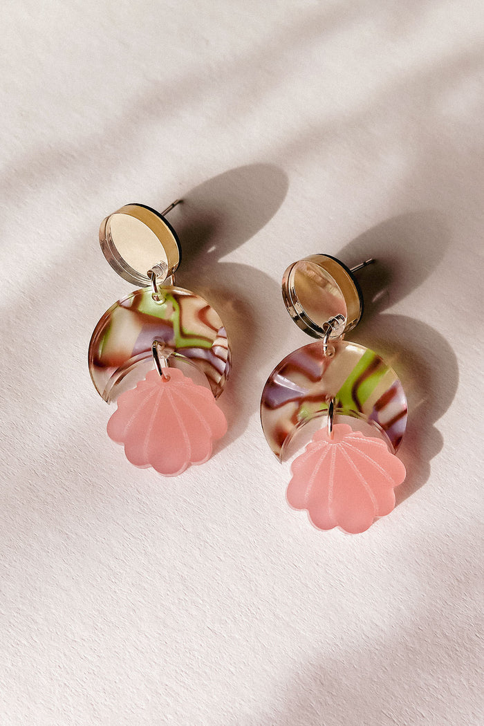 A pair of handmade earrings with round, marbled discs in cream, brown, and green, each featuring a pink, translucent, flower-shaped charm hanging below; lightweight and displayed on a light textured surface with soft shadows.