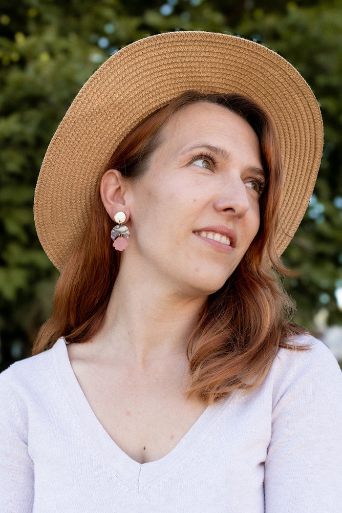 A woman with reddish-brown hair wearing a wide-brimmed straw hat, lightweight handmade seashell earrings, and a light V-neck top looks to the side, smiling softly. A blurred background of green trees is visible.