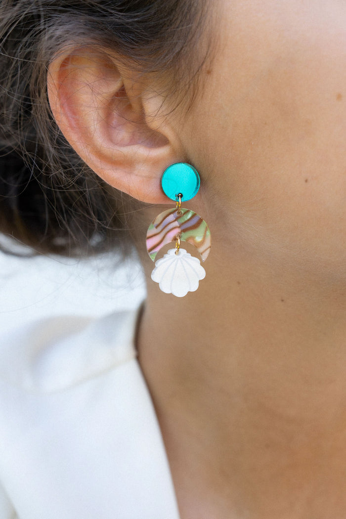 Close-up of a woman’s ear wearing a handmade, colorful earring with a turquoise stud, wavy striped section, and white floral-shaped dangle. Her hair is pulled back and she is wearing a white top. Lightweight for all-day comfort.