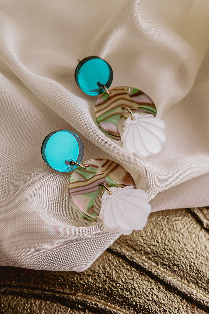 A pair of handmade Seashell Earrings featuring round turquoise studs, marbled circular pieces in green, pink, and clear tones, and lightweight white seashell-shaped charms, displayed on draped white fabric.