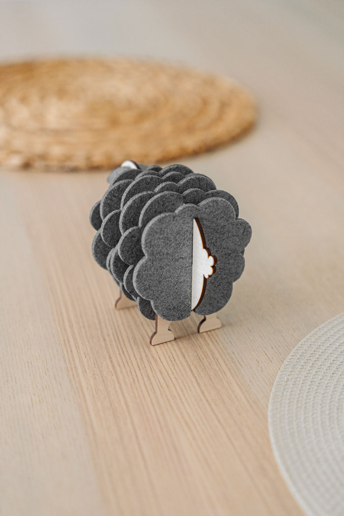 A wooden sheep-shaped coaster holder with dark felt coasters stands on a light table, adding a charming touch to your home decor. Woven placemats are visible in the background.
