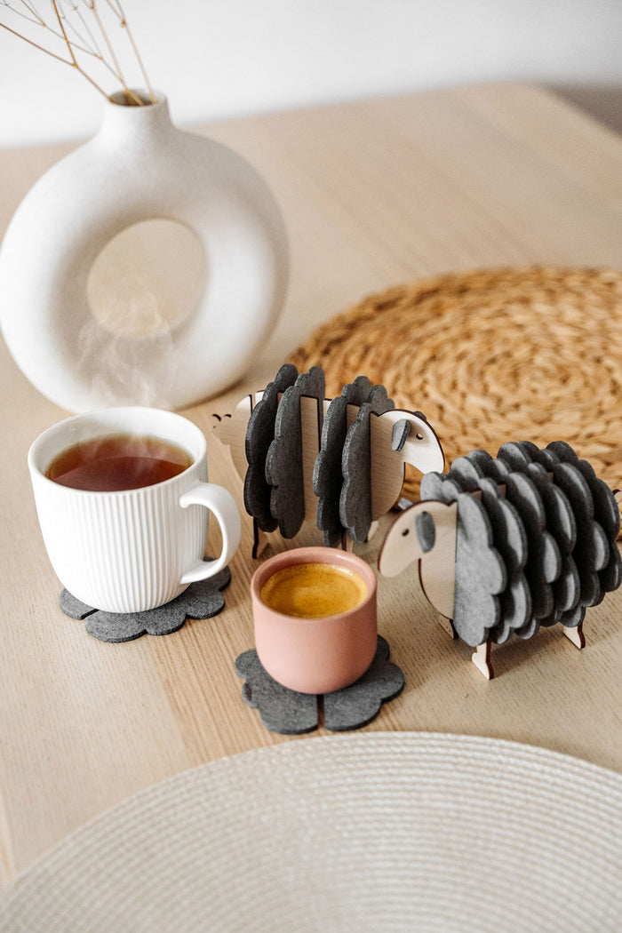 Two wooden sheep-shaped coaster holders with dark gray felt coasters add a touch of charming home decor to a light wooden table, paired with a white mug, pink cup, woven placemat, and modern white vase with dried stems.
