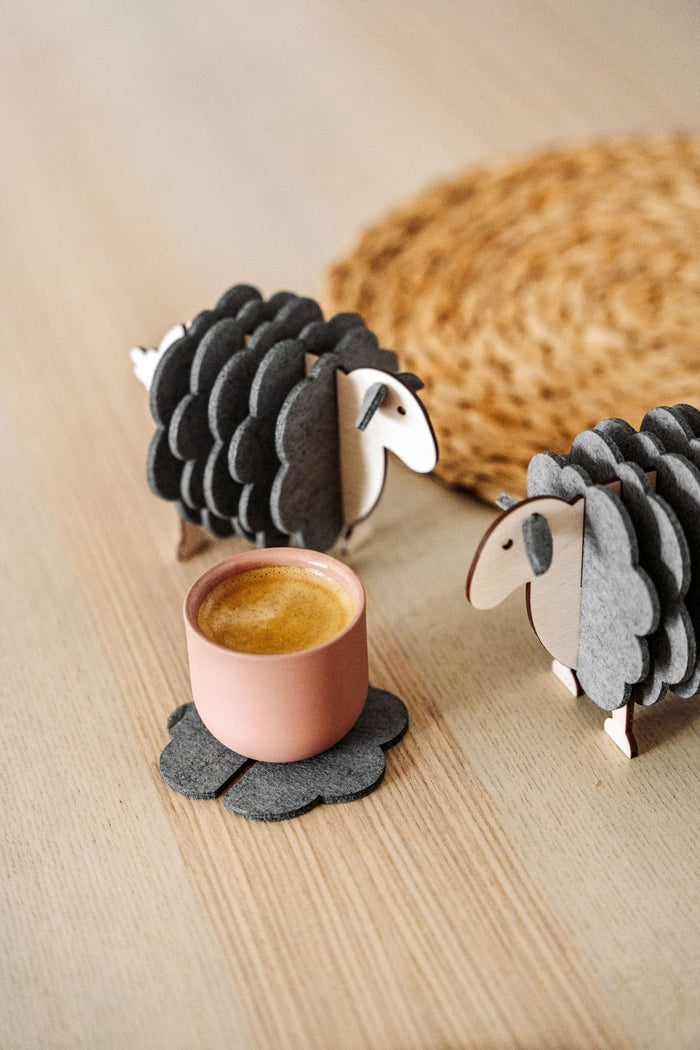 A pink cup rests on a gray, flower-shaped felt coaster beside two wooden sheep figurines—charming home decor—placed on a light wooden surface, with a woven mat partially visible in the background.