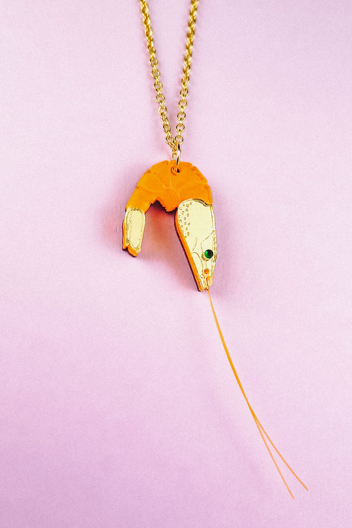 A whimsical Shrimp Pendant Necklace: this gold necklace features a playful orange shrimp pendant with a green gem eye and thin gold whiskers, beautifully displayed on a pastel pink background.