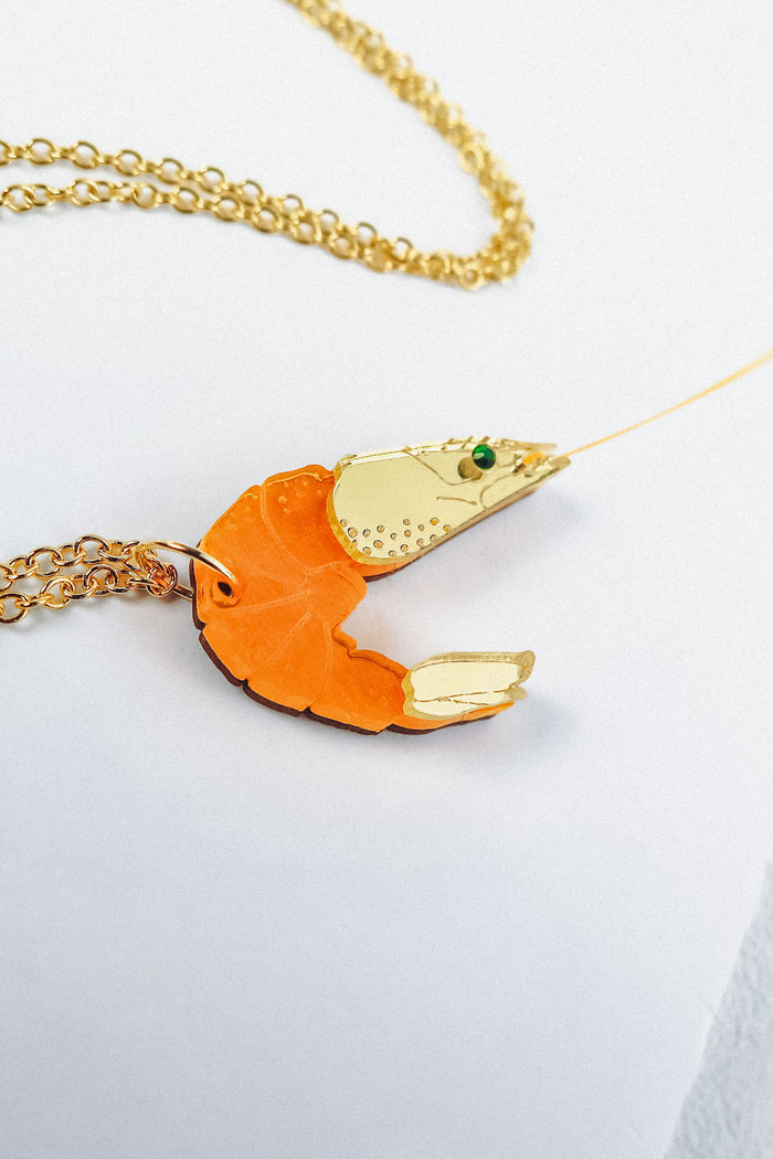 A playful Shrimp Pendant Necklace with an orange body and gold claw details rests on a white surface, showcasing its whimsical design.