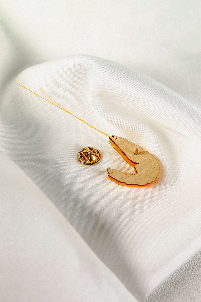 A Shrimp Pin accessory with orange accents and a gold pin is displayed on a soft, white, textured fabric background, adding a playful wardrobe charm to any outfit.