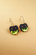 A pair of limited edition Poisoned Apple Earrings shaped like apples with neon green bottoms and black, drippy tops resembling poison, hanging from gold hoops, displayed on a beige background.