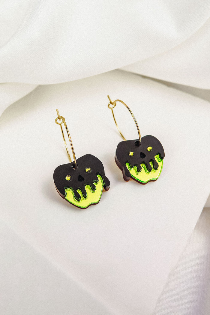A pair of limited edition gold hoop earrings featuring black apple-shaped charms with bright green dripping accents, inspired by a Poisoned Apple Earrings Halloween design, displayed on a white fabric background.