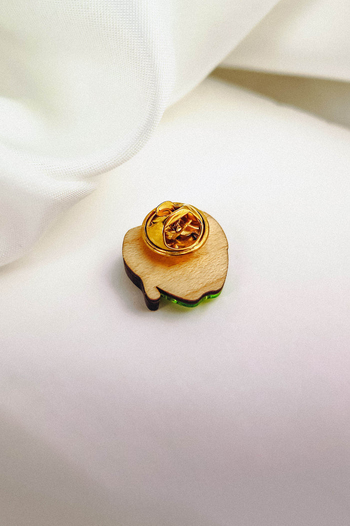 A wooden Poisoned Apple Badge pin with a gold clasp rests on a white fabric background. Perfect for Halloween lovers, its light brown surface features a green edge visible at the bottom. Limited stock available!.