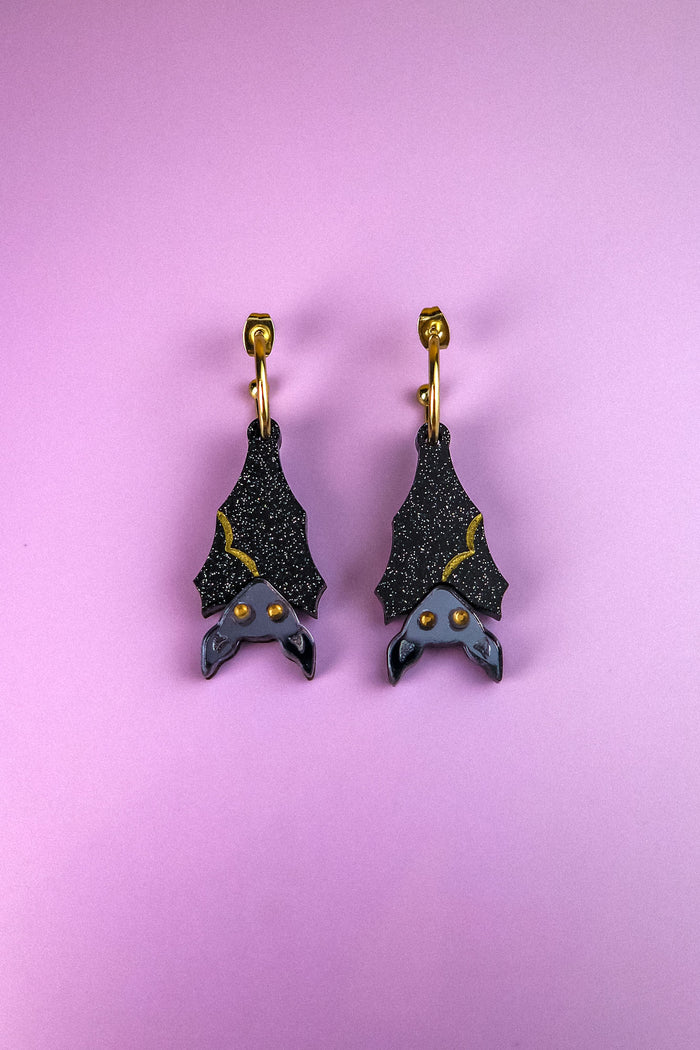A pair of black hand-painted bat-shaped earrings with yellow accents and orange eyes on gold hooks, displayed on a pastel purple background. These allergy-free earrings add a whimsical touch to any outfit.