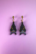A pair of black hand-painted bat-shaped earrings with yellow accents and orange eyes on gold hooks, displayed on a pastel purple background. These allergy-free earrings add a whimsical touch to any outfit.