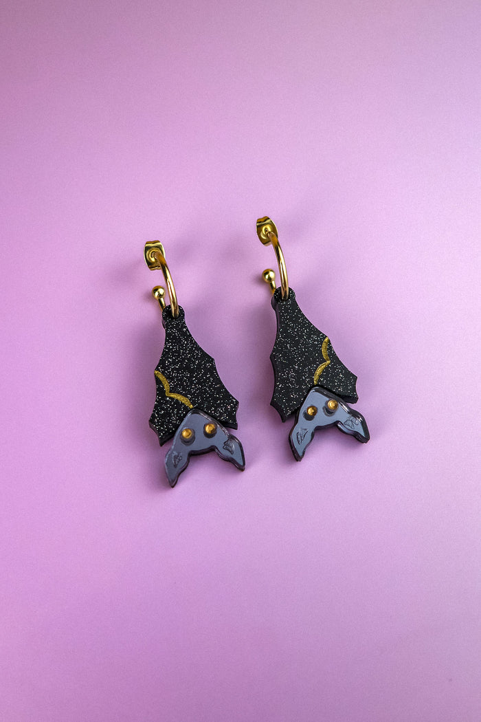 A pair of hand-painted bat-shaped earrings with black, glittery wings and gold accents, hanging from gold hoops, displayed on a pastel purple background. These allergy-free earrings combine unique design with comfortable wear.