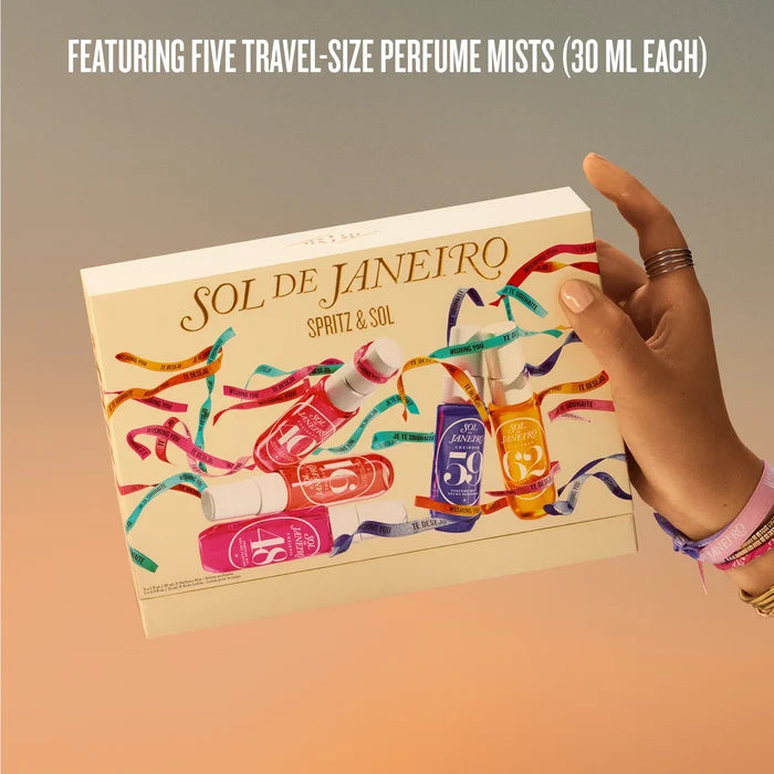 A hand holds a beige box of SOL DE JANEIRO Spritz & Sol travel-size perfume mists against a warm gradient background. The box features colorful illustrations of miniature perfume bottles wrapped in multicolored streamers, with text at the top reading 'FEATURING FIVE TRAVEL-SIZE PERFUME MISTS (30 ML EACH)'.
