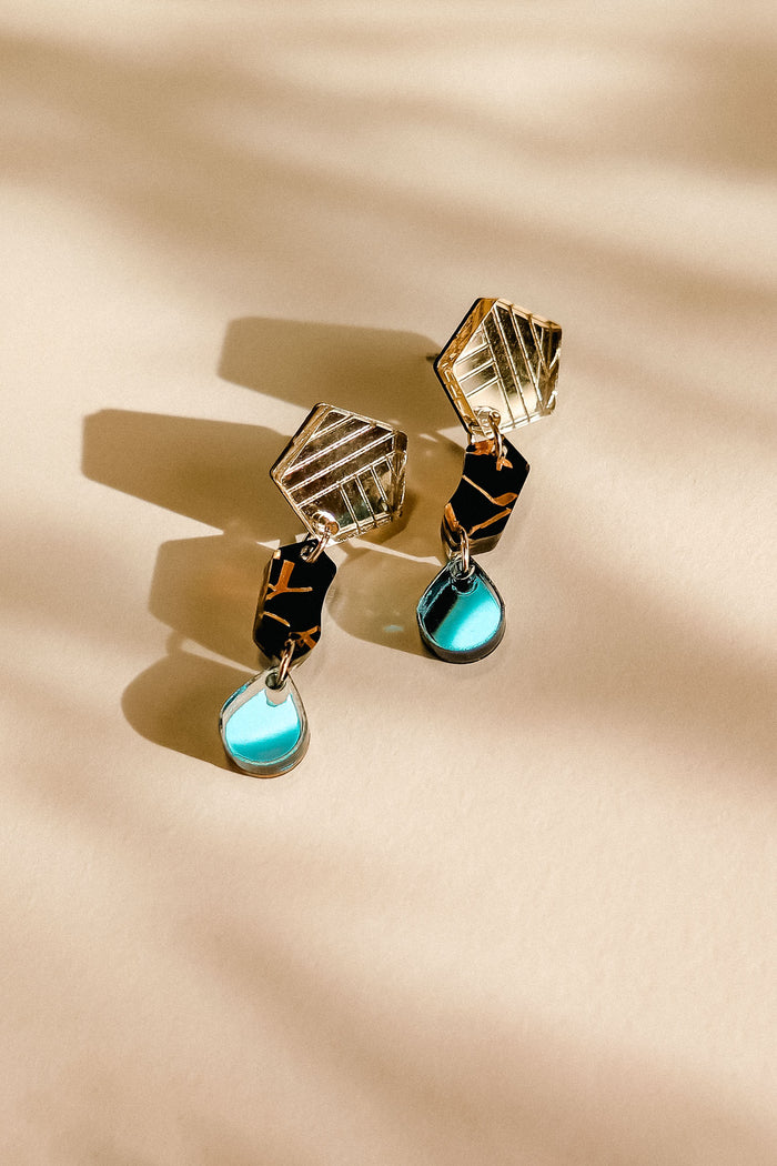 A pair of handcrafted geometric earrings with gold angular tops, black middle sections, and blue tear-drop pendants, displayed on a light beige surface with soft shadow patterns.