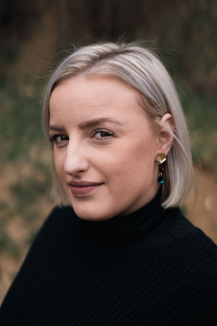A woman with short, straight blonde hair and handcrafted gold earrings looks at the camera with a slight smile, wearing a black turtleneck sweater. The blurred background features eco-friendly greenery and warm brown tones.