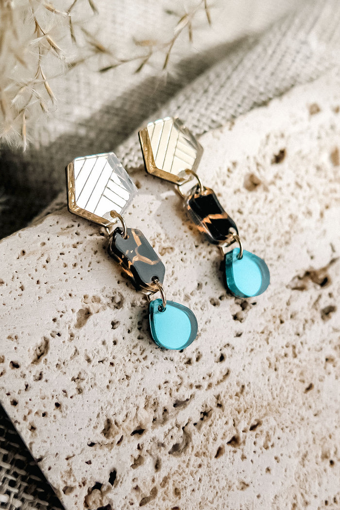 A pair of handcrafted geometric drop earrings with gold, tortoiseshell, and turquoise blue elements rest on a textured stone surface with a neutral fabric and dried plants in the background.