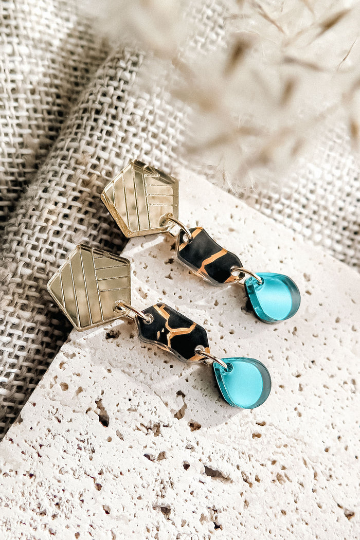 A pair of stylish, handcrafted tear-drop earrings featuring gold hexagonal studs, black and gold patterned middle segments, and translucent blue dangles, displayed on textured fabric and stone surfaces.