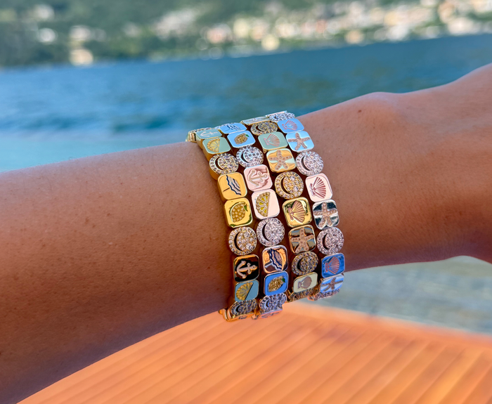 A close-up of a person's wrist wearing three stacked gold-tone bracelets adorned with colorful, engraved Mediterranean-style sea-themed charms, set against a blurred background of blue water and a wooden deck.