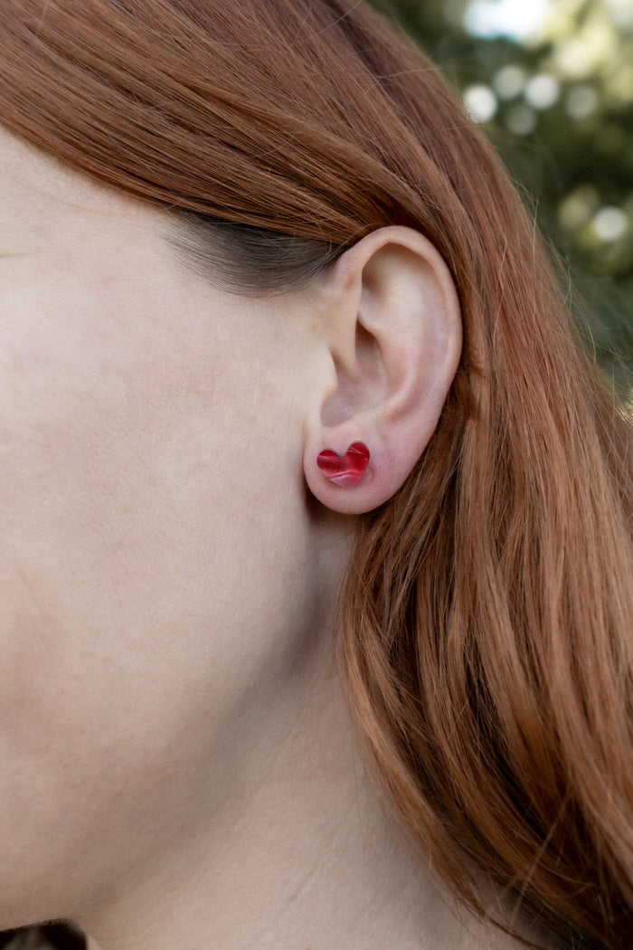 Close-up of a person with long reddish-brown hair wearing a red Tiny Heart Stud in their left ear, highlighting its minimalist charm. The background is blurred greenery.